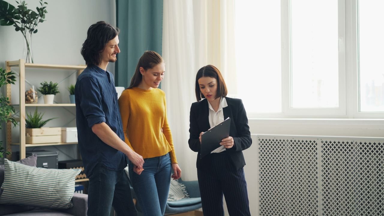 Home A young couple discusses home buying options with a real estate agent inside a well-lit modern room.