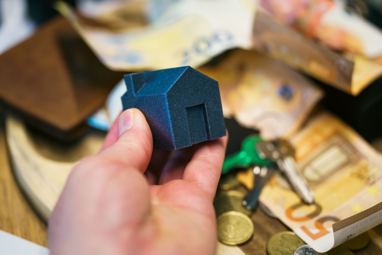 Home Close-up of a hand holding a small house model surrounded by euro banknotes and keys, symbolizing real estate investment.